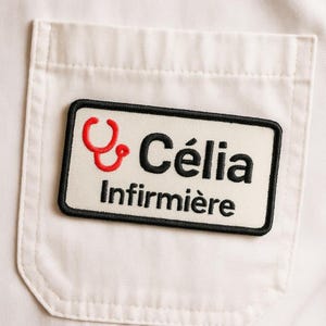 Peut inclure: Un badge blanc avec un bord noir, portant le nom "Célia" en noir et un symbole de stéthoscope rouge. Le mot "Infirmière" est imprimé en dessous en noir. Le badge est fixé sur une poche blanche.