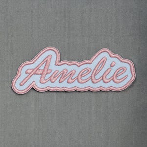 Peut inclure: Écusson brodé bleu clair avec le nom "Amélie" en rose en écriture cursive. Le patch a une bordure rose et est posé sur un fond gris. Parfait pour personnaliser des vêtements ou des accessoires.