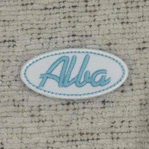 Peut inclure: Un écusson ovale avec le nom "Alba" brodé en bleu clair. Le patch a un fond blanc et une bordure cousue bleu clair. Il est conçu pour être cousu sur des vêtements ou des accessoires.