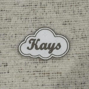 May include: A white cloud-shaped patch with the name "Kays" in a cursive font. The patch has a dark green border and four small star-like designs. The patch is on a textured, off-white background.