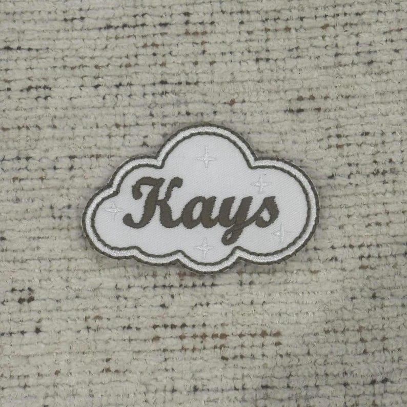 May include: A white cloud-shaped patch with the name "Kays" in a cursive font. The patch has a dark green border and four small star-like designs. The patch is on a textured, off-white background.