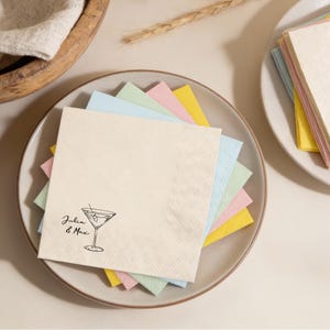 Op de afbeelding: Een stapel pastelkleurige cocktailservetten op een bord. De bovenste servet is crèmekleurig met een zwarte lijntekening van een martiniglas en de tekst "Julia & Max". Andere servetten zijn lichtblauw, groen, roze en geel.