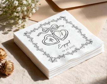 May include: A stack of white paper napkins with a black floral border and two heart-shaped pendants tied with a bow. The pendants have the letters "M" and "A" inside. The word "Engaged" and the date "08.25.2025" are also printed on the napkins.