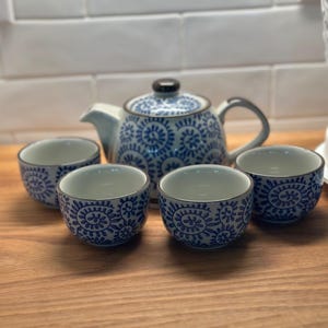May include: A ceramic tea set consisting of a teapot with a lid and three matching tea cups. Each piece features a white base with a repeating blue spiral and leaf pattern. The teapot has a curved handle and spout, and all items have dark rims.