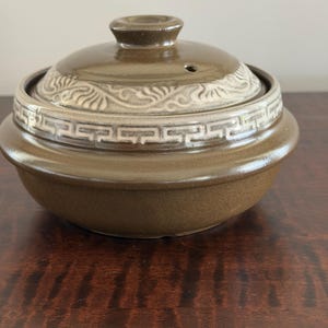 Peut inclure: Un pot de cuisson rond en céramique brune avec un couvercle assorti. Le couvercle présente un bouton central, un petit évent à vapeur et une bande décorative avec un motif grec blanc et des motifs de feuilles tourbillonnantes en relief.