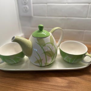 Puede incluir: Un juego de té de cerámica blanca que consta de una tetera con tapa verde y dos tazas a juego, todas decoradas con un patrón de hojas verdes y verde claro. El conjunto está dispuesto sobre una bandeja de servicio ovalada blanca a juego.