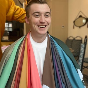 Op de afbeelding: Een persoon glimlacht, omringd door stofstalen in verschillende kleuren, waaronder groen, roze, bruin en blauw. De stalen zijn om de nek van de persoon gedrapeerd en tonen een reeks kleuren voor mode of design.