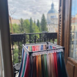 Puede incluir: Un expositor lleno de muestras de tela coloridas. Las muestras están dispuestas en un arcoíris de colores, desde el rosa claro hasta el azul marino oscuro. El expositor está situado cerca de una ventana con vistas a una ciudad y un edificio con cúpula.