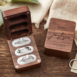 May include: A wooden ring box with three ring slots, open to display three rings. The box is next to a closed wooden box with the inscription "Mr. & Mrs. Jackson 10.12.2025". A silver ring is also visible.