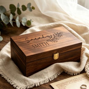 Peut inclure: Une boîte à souvenirs rectangulaire en bois brun foncé avec un fermoir en métal doré. Le couvercle présente un motif floral gravé au laser ainsi que le texte Sarah SMITH. La boîte a une finition en grain de bois lisse et un couvercle à charnière.