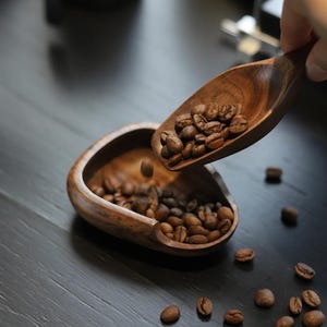 以下が含まれることがあります： 木製のスプーンがコーヒー豆を木製のボウルに注いでいます。スプーンとボウルは濃い色の木でできており、豆は豊かな茶色です。いくつかの豆が暗い表面に散らばっています。