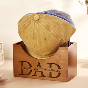 May include: A rectangular wooden storage box with a curved front cutout, featuring the engraved text DAD with the names Emily, Aiden, James, and Grace separated by heart symbols. The box holds a stack of three baseball caps in tan, blue, and grey.