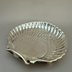 May include: A polished silver-toned metal dish shaped like a scallop shell. The dish features a concave interior with radiating fluted ridges that extend from the hinge area to the scalloped outer edge, creating a reflective and textured surface.