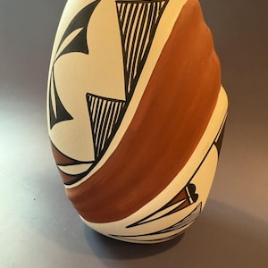 May include: A cream-colored ceramic vase with a rounded shape. The vase features a wide band of reddish-brown paint and is decorated with black geometric designs. The vase has a traditional, handcrafted appearance.