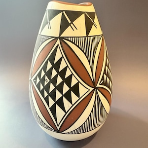 May include: A handcrafted, off-white ceramic vase with a unique, teardrop shape. The vase features intricate geometric patterns in black, brown, and white, including triangles, diamonds, and curved lines. The rim is a warm brown color.