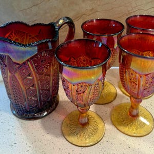 May include: A vintage iridescent glass pitcher and four matching goblets. The set features a rich red hue with intricate embossed patterns and a golden-yellow base. The pitcher has a handle and a flared spout.
