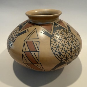 May include: A tan ceramic pot with a rounded body and a short neck. The surface features hand-painted black and terracotta geometric patterns, including circular motifs with grid lines and triangular shapes arranged around the exterior.
