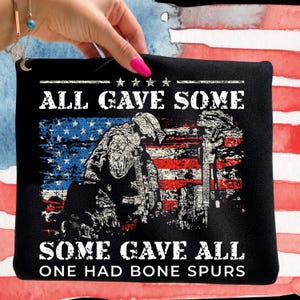 Puede incluir: Una camiseta negra con un gráfico desgastado de un soldado arrodillado ante un rifle y un casco, sobre una bandera estadounidense descolorida. El texto dice ALL GAVE SOME, SOME GAVE ALL, ONE HAD BONE SPURS en una fuente blanca y desgastada.