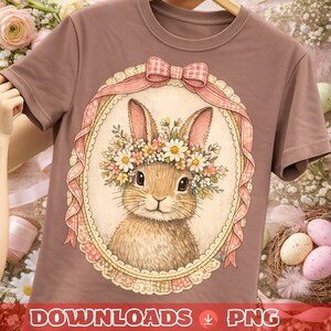 Puede incluir: Camiseta marrón con una ilustración de un conejito con una corona de flores, enmarcada por un óvalo decorativo con una cinta y un lazo rosas. El texto "DOWNLOADS PNG" está en la parte inferior. La camiseta está colgada en una percha de madera.