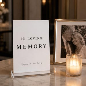 May include: A white sign with the words "IN LOVING MEMORY" and "Forever in our hearts" in black text. A framed black and white photo and a lit candle in a glass holder are also visible.