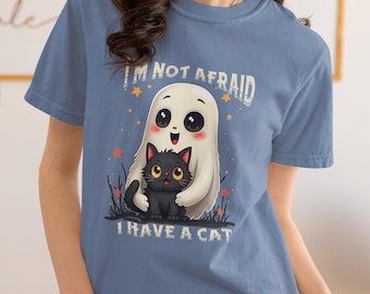 May include: A blue t-shirt featuring a cartoon ghost hugging a black cat. The text "I'M NOT AFRAID I HAVE A CAT" is printed above and below the image. The design includes stars and grass.