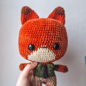 Puede incluir: Una muñeca de zorro de ganchillo hecha a mano con un cuerpo naranja afelpado y texturizado y orejas con puntas marrones. El zorro tiene grandes ojos negros, un hocico blanco y lleva una chaqueta verde con una corbata azul oscuro. La muñeca es sostenida por una mano.