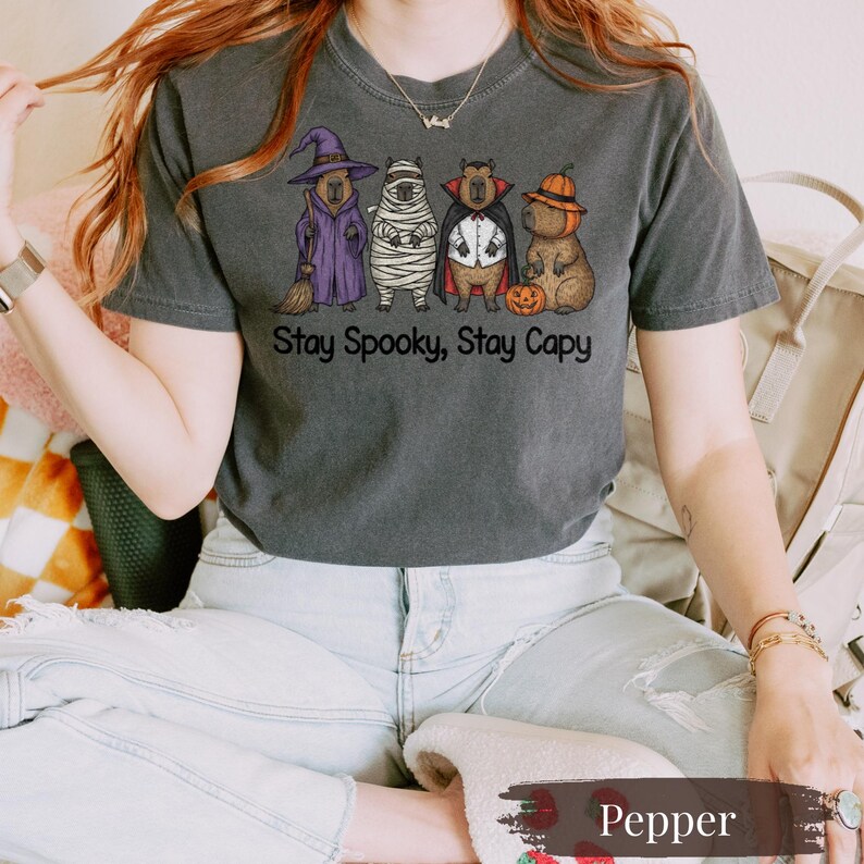 Pepper Comfort Colors shirt featuring a quirky Halloween capybara design with four dressed-up rodents: witch, mummy, vampire, and pumpkin. Ideal spooky shirt for fans of funny animal memes, zoo animals, and rodent lovers. Soft, oversized tee for Halloween parties, trick-or-treat night, or fall photoshoots. Trendy pet lover gift that blends spooky fun with cozy capybara energy in muted charcoal tones.