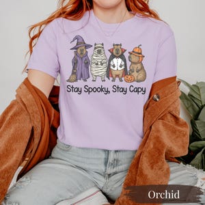 Cute Halloween animal shirt on orchid Comfort Colors tee featuring dressed-up capybaras: witch, mummy, vampire, and pumpkin. Funny Halloween aesthetic shirt for capybara lovers, pet parents, zoo goers, and quirky fall fashion fans. Aesthetic purple tee for spooky season with vintage comfort fit. Great gift for capybara meme fans and Halloween lovers who want a soft, stylish and fun seasonal top.