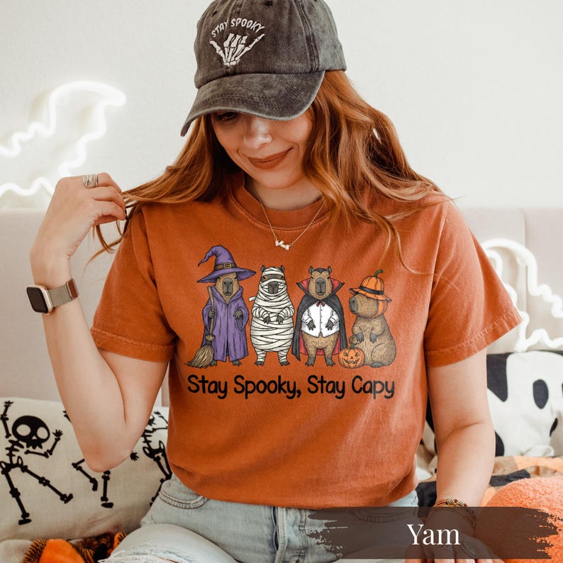 Spooky season capybara shirt printed on a vibrant yam Comfort Colors tee. Design shows four funny Halloween capybaras in witch, mummy, vampire, and pumpkin costumes. Unique fall shirt for animal lovers, Halloween party goers, and pet enthusiasts. Bold and playful Halloween outfit idea with a vintage-washed feel. Perfect for zoo fans, capybara moms, and anyone who loves weird cute animals in cozy fall colors.