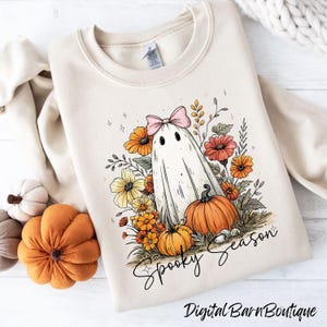 May include: Cream-colored sweatshirt featuring a whimsical ghost design with a pink bow, surrounded by flowers and pumpkins. The words "Spooky Season" are written in a script font. The design is a watercolor style, perfect for Halloween.