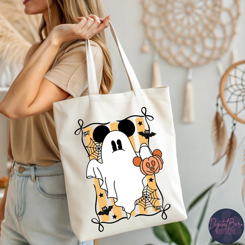 May include: A cream-colored tote bag featuring a Halloween-themed design. The design includes a white ghost with Mickey Mouse ears, a jack-o'-lantern, bats, and spiderwebs, set against a yellow and orange striped background. The bag has white handles.