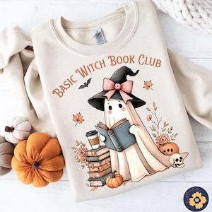 May include: Beige sweatshirt with the text "Basic Witch Book Club" and a cartoon ghost wearing a witch hat and reading a book. The design includes fall-themed elements like pumpkins, leaves, and flowers. A bat graphic is also included.