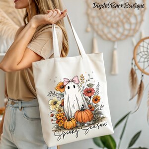 May include: Cream-colored tote bag featuring a ghost with a pink bow, surrounded by orange and yellow flowers, and pumpkins. The words "Spooky Season" are written in a script font. The bag is held by a person.