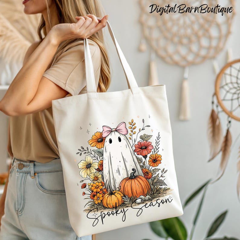 May include: Cream-colored tote bag featuring a ghost with a pink bow, surrounded by orange and yellow flowers, and pumpkins. The words "Spooky Season" are written in a script font. The bag is held by a person.
