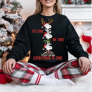 Puede incluir: Sudadera negra con personajes de Snoopy con gorros de Papá Noel y cuernos de reno, con el texto "It's Fine, I'm Fine, Everything is Fine" en rojo. La persona lleva un pantalón de pijama a cuadros rojos y blancos.