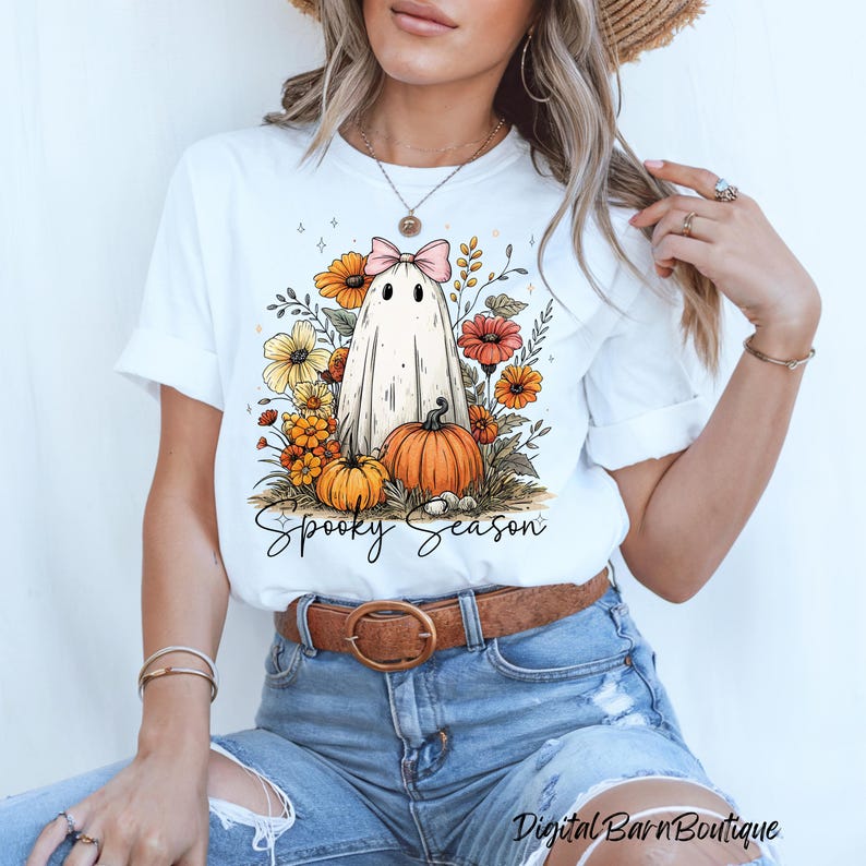 May include: White t-shirt featuring a cartoon ghost with a pink bow, surrounded by flowers and pumpkins. The words "Spooky Season" are written in a script font below the design. The shirt is paired with blue jeans and a brown belt.