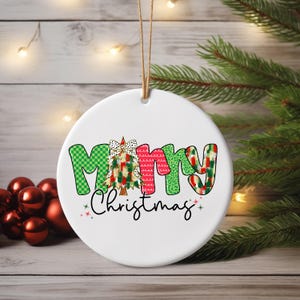 Op de afbeelding: Een witte, ronde kerstversiering met de tekst "Merry Christmas" in een kleurrijk, patroonontwerp. De letter "M" is groen met een geruit patroon, de "R" is rood en de "Y" is groen met een feestelijk patroon. Een kerstboom staat in het midden.