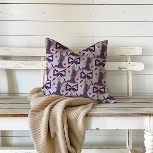 May include: A square cushion featuring a repeating pattern of a cartoon queen, bows, and apples on a light purple background. The cushion is on a white bench with a beige blanket. The pattern is in shades of purple and red.