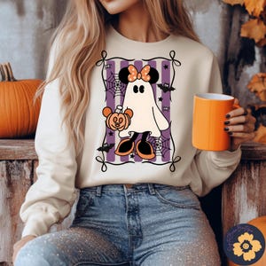 May include: Cream-colored sweatshirt featuring a cartoon ghost holding a pumpkin. The ghost has a bow and mouse ears. The design includes purple stripes, spiderwebs, and bats. An orange mug is held in the hand.