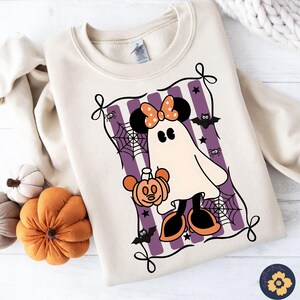 May include: A cream-colored sweatshirt featuring a Halloween-themed graphic. The graphic depicts a ghost in the shape of Minnie Mouse, holding a pumpkin. The design includes purple stripes, spiderwebs, bats, and stars. The sweatshirt has a crew neck and long sleeves.