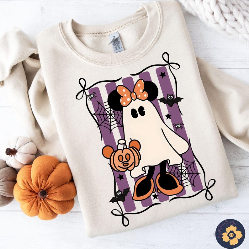 May include: A cream-colored sweatshirt featuring a Halloween-themed graphic. The graphic depicts a ghost in the shape of Minnie Mouse, holding a pumpkin. The design includes purple stripes, spiderwebs, bats, and stars. The sweatshirt has a crew neck and long sleeves.