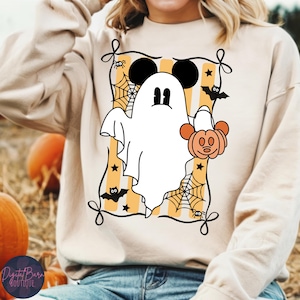 May include: A beige sweatshirt featuring a Halloween-themed graphic. The design includes a white ghost with Mickey Mouse ears, holding a pumpkin, set against a yellow and white striped background with bats, spiders, and spiderwebs. The sweatshirt has a crew neck.