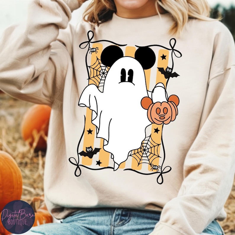 May include: A beige sweatshirt featuring a Halloween-themed graphic. The design includes a white ghost with Mickey Mouse ears, holding a pumpkin, set against a yellow and white striped background with bats, spiders, and spiderwebs. The sweatshirt has a crew neck.