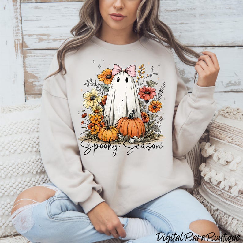 May include: Beige sweatshirt featuring a whimsical ghost design with a pink bow, surrounded by colorful flowers and pumpkins. The words "Spooky Season" are written below the graphic. A seasonal, festive design.