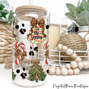 May include: A clear glass tumbler with a bamboo lid, decorated with Christmas-themed graphics. The design includes a bell with the text "Tis the Season", a candy cane, and a Christmas tree, all adorned with leopard print bows and accents.
