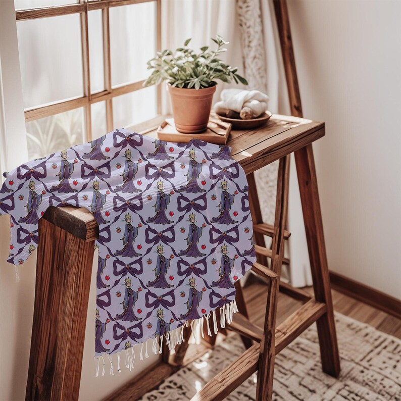 May include: A light purple scarf featuring a cartoon queen holding an apple, bows, and red apples. The scarf has a white fringe and is draped over a wooden surface.