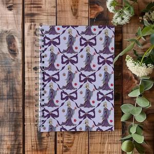 May include: Spiral-bound notebook with a repeating pattern of a queen holding an apple, bows, and apples on a light purple background. The queen wears a purple gown and a gold crown. The notebook is on a wooden surface.