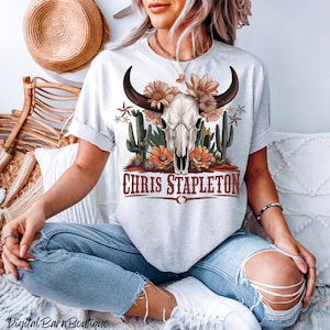 May include: A light gray t-shirt featuring a skull with horns, flowers, and cacti. The text "CHRIS STAPLETON" is printed in a brown, western-style font. The shirt is worn with blue jeans.