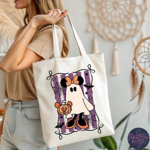 May include: A cream-colored tote bag featuring a Halloween-themed graphic. The design includes a ghost character with a Minnie Mouse bow, holding a pumpkin, set against purple stripes, bats, and spiderwebs. The bag has long handles.