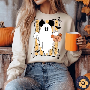 May include: Cream-colored sweatshirt featuring a Halloween-themed graphic. The design includes a white ghost with Mickey Mouse ears, holding a pumpkin, set against a yellow and orange striped background with spiderwebs and bats. An orange mug is held in the hand.